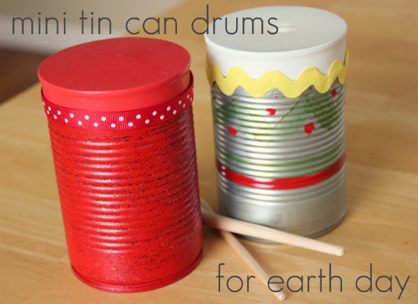 Two decorated mini tin can drums are shown. One is painted red with a lid, and the other has a zig-zag yellow border with painted designs. Wooden sticks lie beside them. The text reads, mini tin can drums for Earth Day.